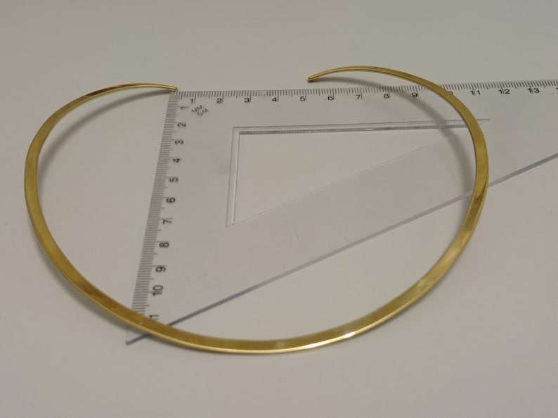 May include: A gold-colored choker necklace, approximately 13 inches in diameter, is displayed on a white surface. The necklace is measured with a clear ruler. The choker has a simple, elegant design.