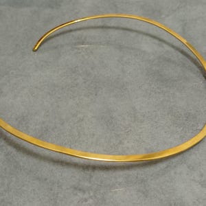 May include: A gold-colored choker necklace with a minimalist design. The necklace is a simple, open-ended band that curves around the neck. The metal has a polished, reflective surface. The jewelry is displayed on a gray surface.