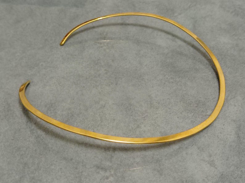 May include: A gold-colored choker necklace with a minimalist design. The necklace is a simple, open-ended band that curves around the neck. The metal has a polished, reflective surface. The jewelry is displayed on a gray surface.
