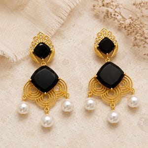 May include: A pair of gold earrings featuring black square gemstones and white pearl drops. The earrings have an ornate, filigree design. The background is neutral. These earrings are a striking jewellery item.