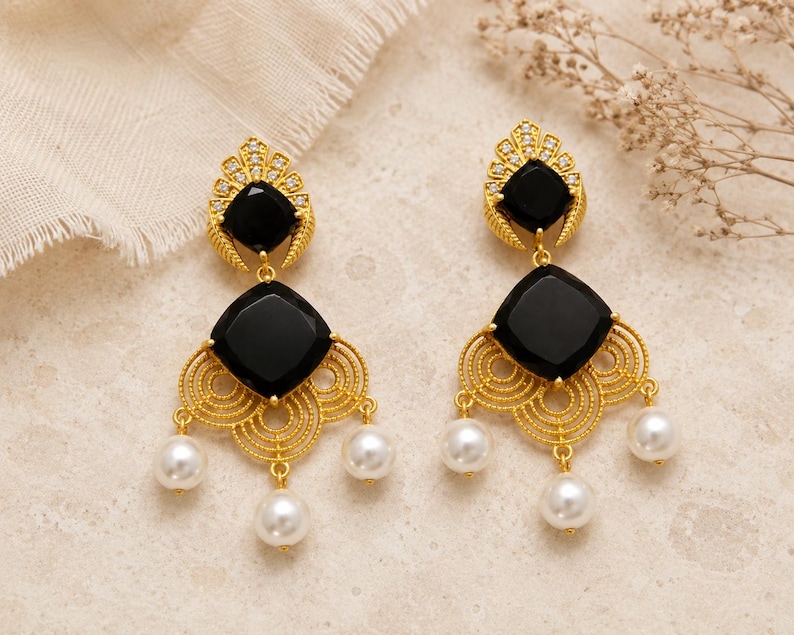 May include: A pair of gold earrings featuring black square gemstones and white pearl drops. The earrings have an ornate, filigree design. The background is neutral. These earrings are a striking jewellery item.