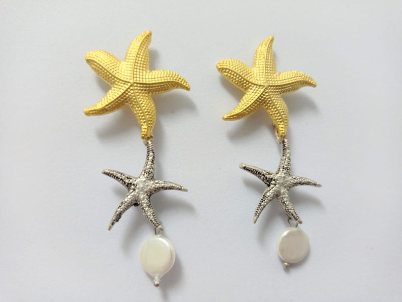 May include: A pair of drop earrings featuring a gold-toned starfish at the top, a silver-toned starfish suspended below, and a small, round, white pearl-like bead dangling at the very bottom of each earring, showcasing a textured surface.