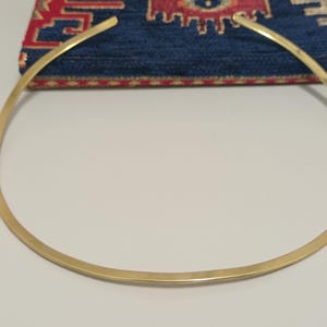 May include: A gold-colored choker necklace with a simple, minimalist design. The necklace is a smooth, curved band. The background features a blue textile with a red and gold pattern.
