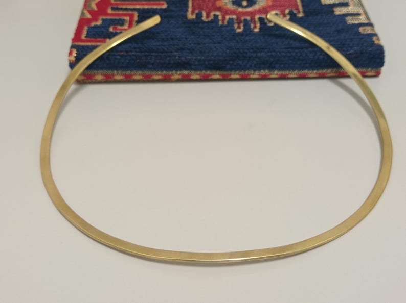 May include: A gold-colored choker necklace with a simple, minimalist design. The necklace is a smooth, curved band. The background features a blue textile with a red and gold pattern.