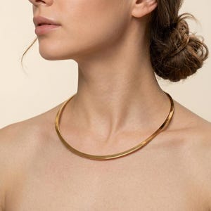 May include: A gold-colored, minimalist collar necklace. The necklace is a simple, curved band that sits close to the neck. The jewelry is a bright, shiny gold color.