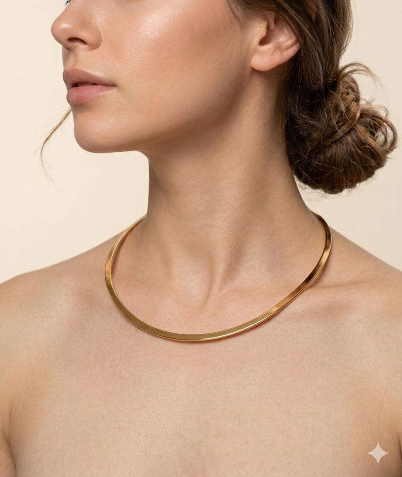 May include: A gold-colored, minimalist collar necklace. The necklace is a simple, curved band that sits close to the neck. The jewelry is a bright, shiny gold color.