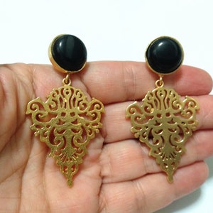 May include: A pair of gold-colored filigree earrings with a black, round stone at the top. The earrings have an ornate, openwork design and are suspended from a gold-colored post.