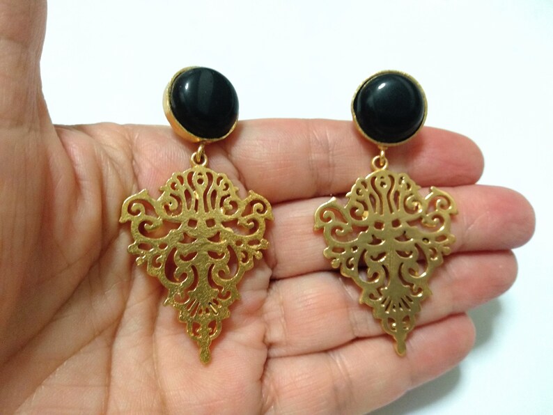 May include: A pair of gold-colored filigree earrings with a black, round stone at the top. The earrings have an ornate, openwork design and are suspended from a gold-colored post.