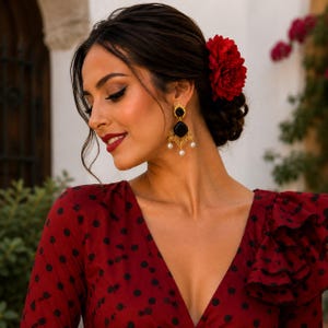 May include: A woman wearing gold earrings with black onyx stones and dangling pearls. The earrings are part of a flamenco-style ensemble, complemented by a red dress with black polka dots and a red flower in her hair. The earrings have a filigree design.