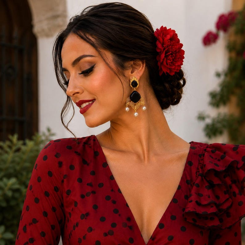 May include: A woman wearing gold earrings with black onyx stones and dangling pearls. The earrings are part of a flamenco-style ensemble, complemented by a red dress with black polka dots and a red flower in her hair. The earrings have a filigree design.