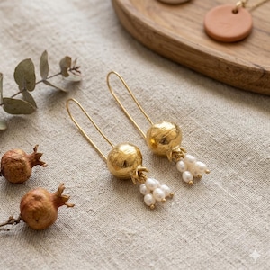 May include: A pair of gold-colored earrings with a pomegranate design, featuring a cluster of small white pearls. The earrings are on a beige linen surface, with dried pomegranates and eucalyptus leaves nearby. A wooden tray with a pendant is in the background.