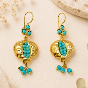 May include: A pair of gold-colored earrings featuring a pomegranate design. The earrings have turquoise-colored beads and a textured surface. The earrings are designed with a hook closure.