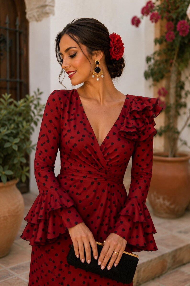 May include: A red polka dot dress with long sleeves and a ruffled shoulder. The dress features a V-neck and flared sleeves. A black clutch and gold earrings are visible. A red flower adorns the hair.