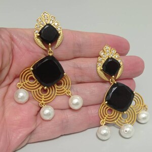 May include: Gold chandelier earrings with black square stones and faux pearl drops. The earrings have a decorative filigree design and are embellished with small crystals. A glamorous accessory for a special occasion.