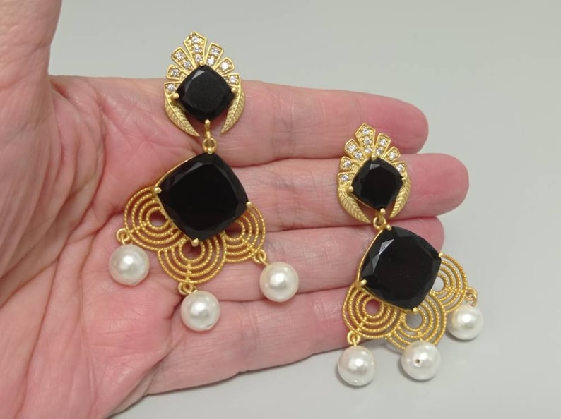 May include: Gold chandelier earrings with black square stones and faux pearl drops. The earrings have a decorative filigree design and are embellished with small crystals. A glamorous accessory for a special occasion.