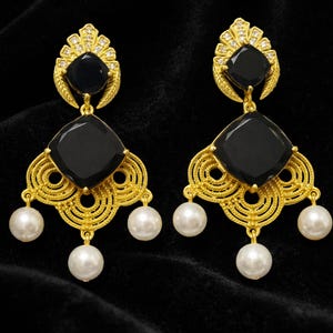 May include: A pair of gold-coloured drop earrings featuring black square-cut stones and white pearl accents. The earrings have an ornate, filigree design with a fan-shaped top and three dangling pearls.