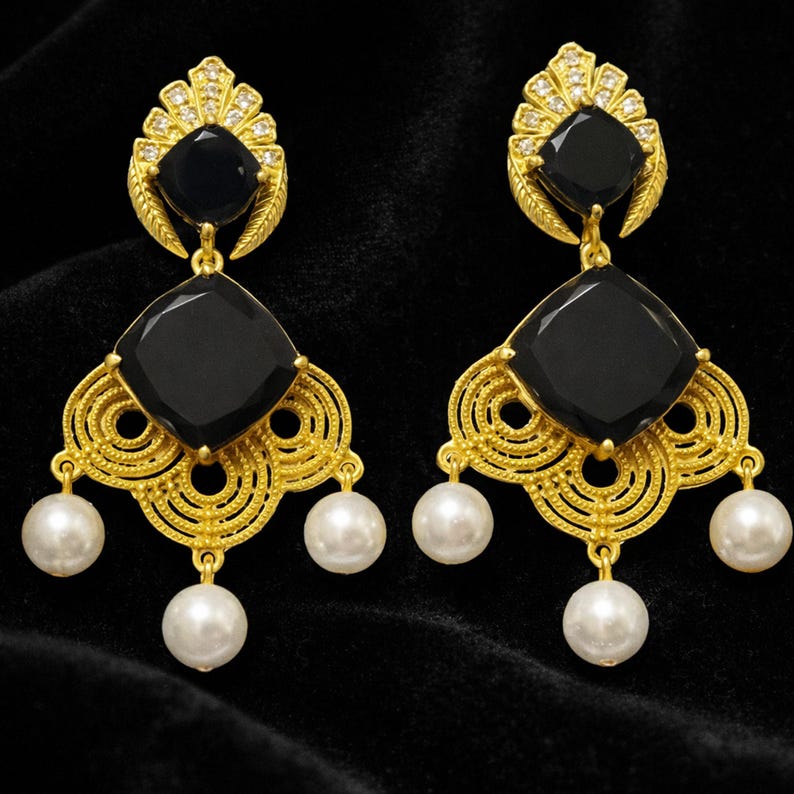 May include: A pair of gold-coloured drop earrings featuring black square-cut stones and white pearl accents. The earrings have an ornate, filigree design with a fan-shaped top and three dangling pearls.