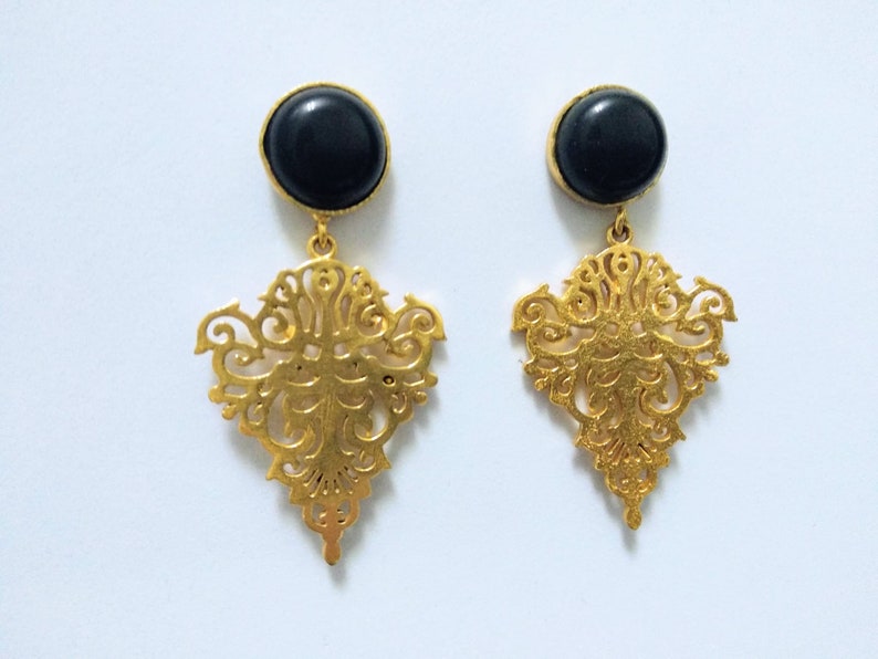 May include: A pair of gold-colored drop earrings. Each earring features a black, circular stone at the top, connected to an ornate, gold filigree design. The filigree has a symmetrical, floral-inspired pattern.