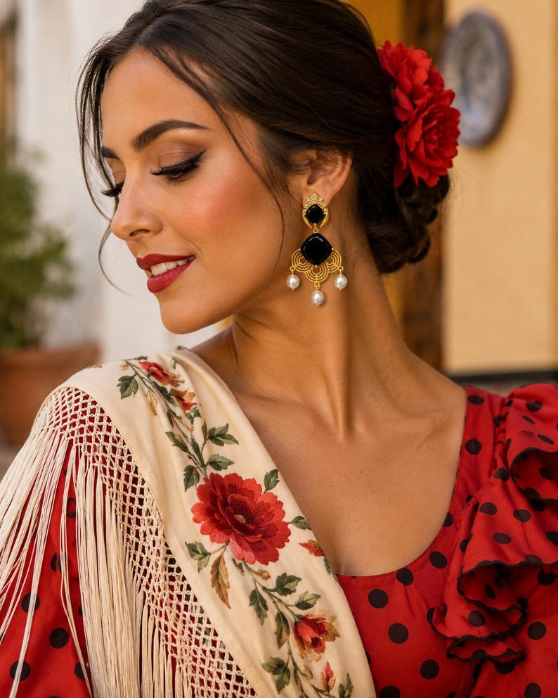 May include: A woman wearing gold earrings with black and pearl details, a red flower in her hair, and a red dress with black polka dots. She is also wearing a cream shawl with red floral embroidery and fringe.