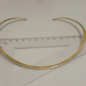 May include: A gold-colored choker necklace with a curved design. The necklace is approximately 14 inches in length and has a simple, elegant style. The necklace is placed on a white surface with a ruler.