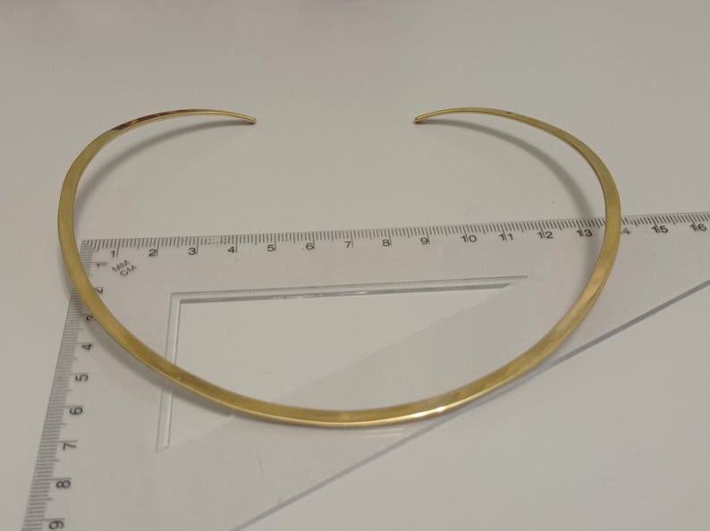 May include: A gold-colored choker necklace with a curved design. The necklace is approximately 14 inches in length and has a simple, elegant style. The necklace is placed on a white surface with a ruler.