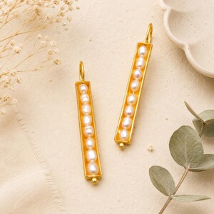 May include: A pair of gold-colored, rectangular drop earrings. Each earring features a row of white pearls set within a gold frame. The earrings are displayed on a cream-colored surface, with a sprig of greenery and a decorative dish in the background.