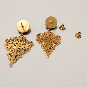 May include: Gold-colored filigree earrings with a detailed, openwork design. The earrings have a post-back closure and a circular backing. The intricate, symmetrical pattern is visible in the dangling portion of the earrings.