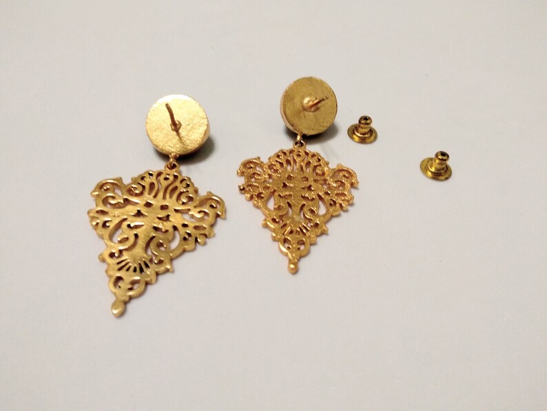 May include: Gold-colored filigree earrings with a detailed, openwork design. The earrings have a post-back closure and a circular backing. The intricate, symmetrical pattern is visible in the dangling portion of the earrings.