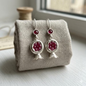 Può includere: Orecchini in argento con design a melograno. Ogni orecchino presenta una piccola gemma rossa nella parte superiore e un gruppo di gemme rosse all'interno di una forma di melograno argentata. Gli orecchini sono esposti su un tessuto grigio.