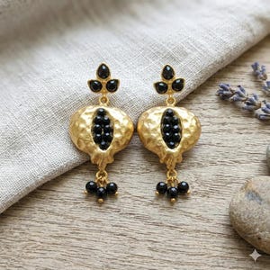 May include: A pair of gold-colored earrings with a pomegranate design. Each earring features a textured, oval-shaped body filled with black beads, topped with three black beads, and dangling black beads at the bottom. The earrings are displayed on a wooden surface.