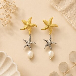 May include: A pair of drop earrings featuring a gold-toned textured starfish at the top, connected to a smaller silver-toned textured starfish below, and finished with a single white pearl dangling at the bottom of each earring.