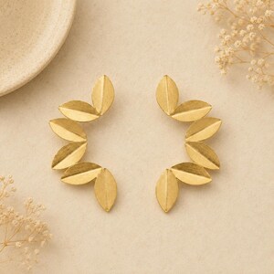May include: A pair of gold-colored leaf-shaped earrings. Each earring features a series of six stylized leaves arranged in a semi-circular pattern. The earrings are displayed on a neutral background, with a ceramic plate and dried flowers.