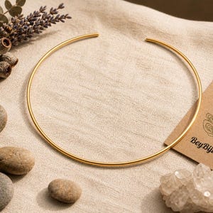 May include: A gold-colored choker necklace with a simple, open design. The jewelry is displayed on a neutral-toned fabric, alongside small stones, dried flowers, and a business card with a peacock logo and the text "Bey Biju Jewelry."