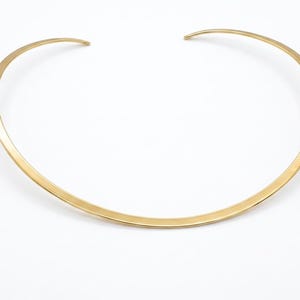 May include: A gold-colored collar necklace with a simple, minimalist design. The necklace has a curved shape that sits around the neck. The metal has a polished, reflective surface, and the necklace is set against a white background.