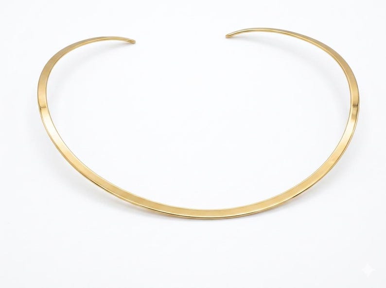 May include: A gold-colored collar necklace with a simple, minimalist design. The necklace has a curved shape that sits around the neck. The metal has a polished, reflective surface, and the necklace is set against a white background.