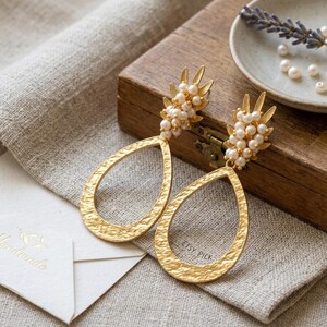 May include: Gold-toned drop earrings with a textured finish and pearl accents. The earrings have a teardrop shape and are displayed on a linen cloth next to a wooden box and a small dish with pearls. The earrings are labeled "ETSY PICK".
