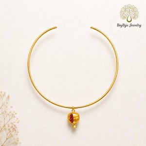 May include: A gold-colored choker necklace with a pomegranate pendant. The pendant is gold with a red gemstone center. The necklace is open at the back and the brand name "BeyBiju Jewelry" is in the upper right corner.