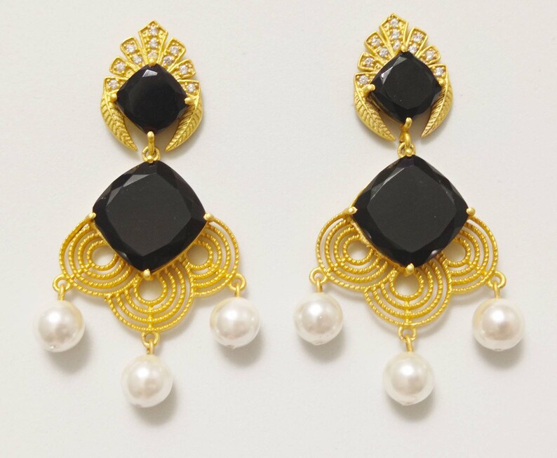 May include: A pair of gold-coloured earrings featuring black square gemstones and white pearl drops. The earrings have a decorative filigree design and are embellished with small clear crystals.