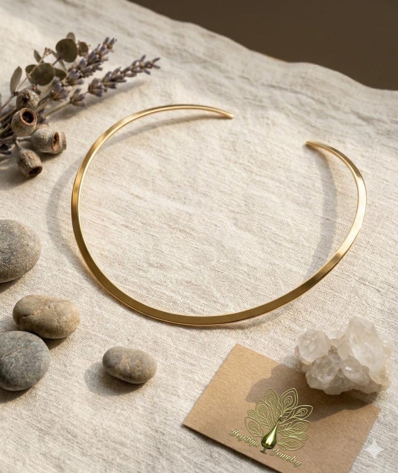 May include: A gold-colored collar necklace with a minimalist design. The necklace is a simple, curved band. The image also includes small stones, dried flowers, and a business card with a peacock logo.