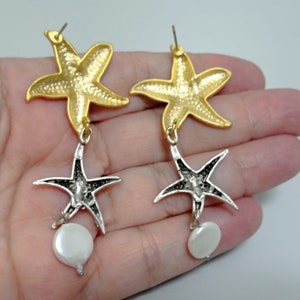 May include: A pair of drop earrings featuring a gold-toned starfish at the top, connected to a silver-toned starfish below, and finished with a small, round, white, iridescent disc-shaped bead dangling from the bottom of each silver starfish.
