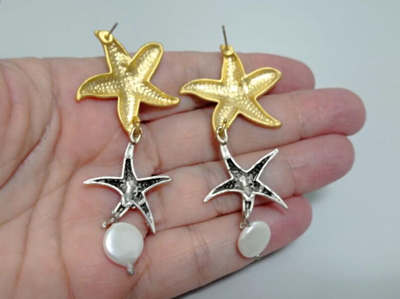 May include: A pair of drop earrings featuring a gold-toned starfish at the top, connected to a silver-toned starfish below, and finished with a small, round, white, iridescent disc-shaped bead dangling from the bottom of each silver starfish.
