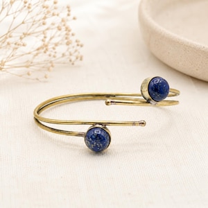 Puede incluir: Una pulsera de puño de metal dorado con dos piedras preciosas esféricas de color azul oscuro. La pulsera tiene un diseño sencillo y abierto con las piedras preciosas engastadas en cada extremo. Las piedras preciosas tienen un aspecto moteado.