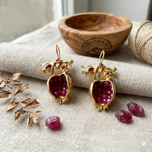 May include: Gold-coloured pomegranate earrings with a deep red interior and small red details. The earrings are presented on a neutral linen surface, with loose gemstones and a wooden bowl in the background.