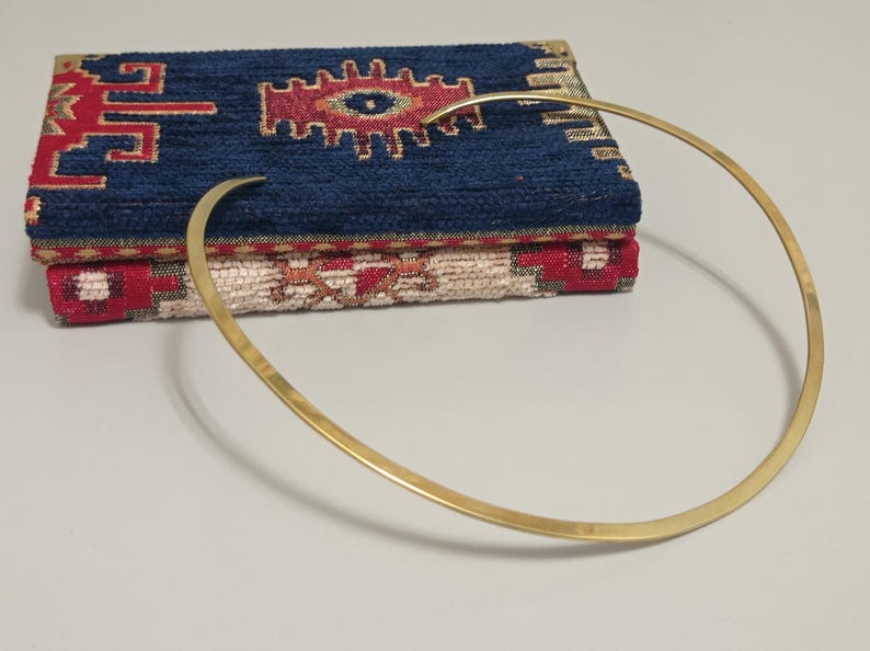 May include: A gold-colored, minimalist collar necklace with a curved design. The necklace rests in front of a stack of two decorative fabric items with a blue and red pattern.