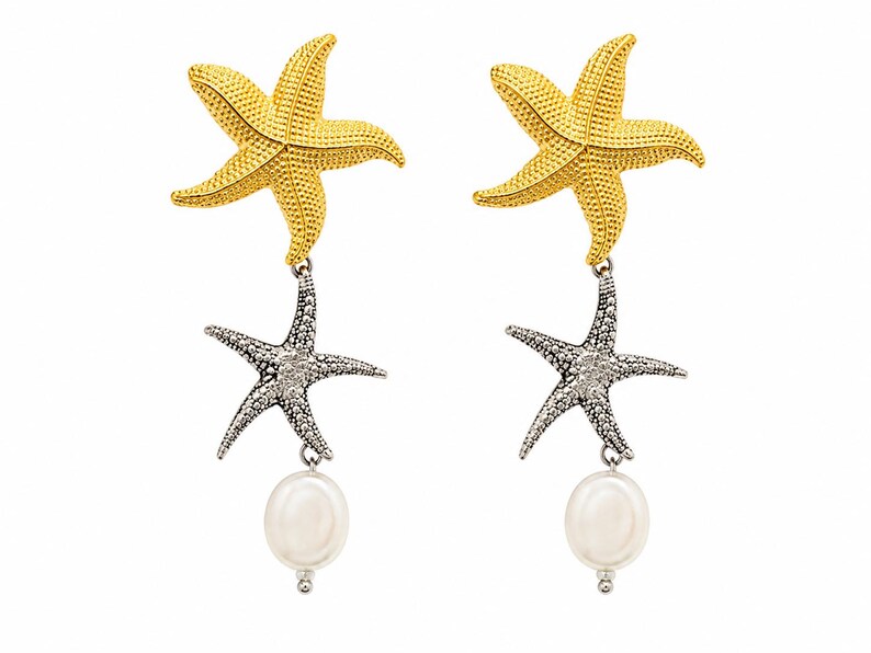 May include: A pair of drop earrings featuring a gold-toned starfish at the top, a silver-toned starfish suspended below, and a single white oval pearl dangling at the bottom. The starfish have a textured, granular surface detail throughout.