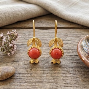 May include: A pair of gold-colored earrings with a red, round stone center. The earrings have a leaf design at the top and a floral base. The earrings are displayed on a wooden surface.