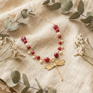 May include: A gold-colored necklace with a dragonfly pendant. The necklace features red and white beads, with a gold chain. The dragonfly pendant is detailed with etched wings. The necklace is displayed on a beige fabric.