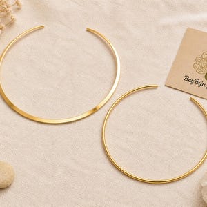 May include: Two gold-colored necklaces with open ends are displayed on a beige surface. One necklace is a flat, wide band, and the other is a thin, round wire. A small card with the text "BeyBiju Jewelry" is also visible.