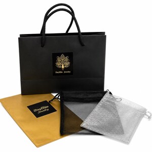 May include: A black shopping bag with a gold logo and black rope handles. The bag is next to a gold envelope, a black mesh bag, and a silver mesh bag. The logo on the bag and envelope reads "DayEllis Jewelry".