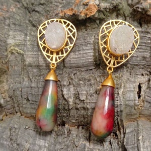 Könnte beinhalten: Goldfarbene Ohrringe mit einem weißen Stein, der in einem geometrischen Design gefasst ist, und einem tropfenförmigen Stein in Rot-, Grün- und Weißtönen.